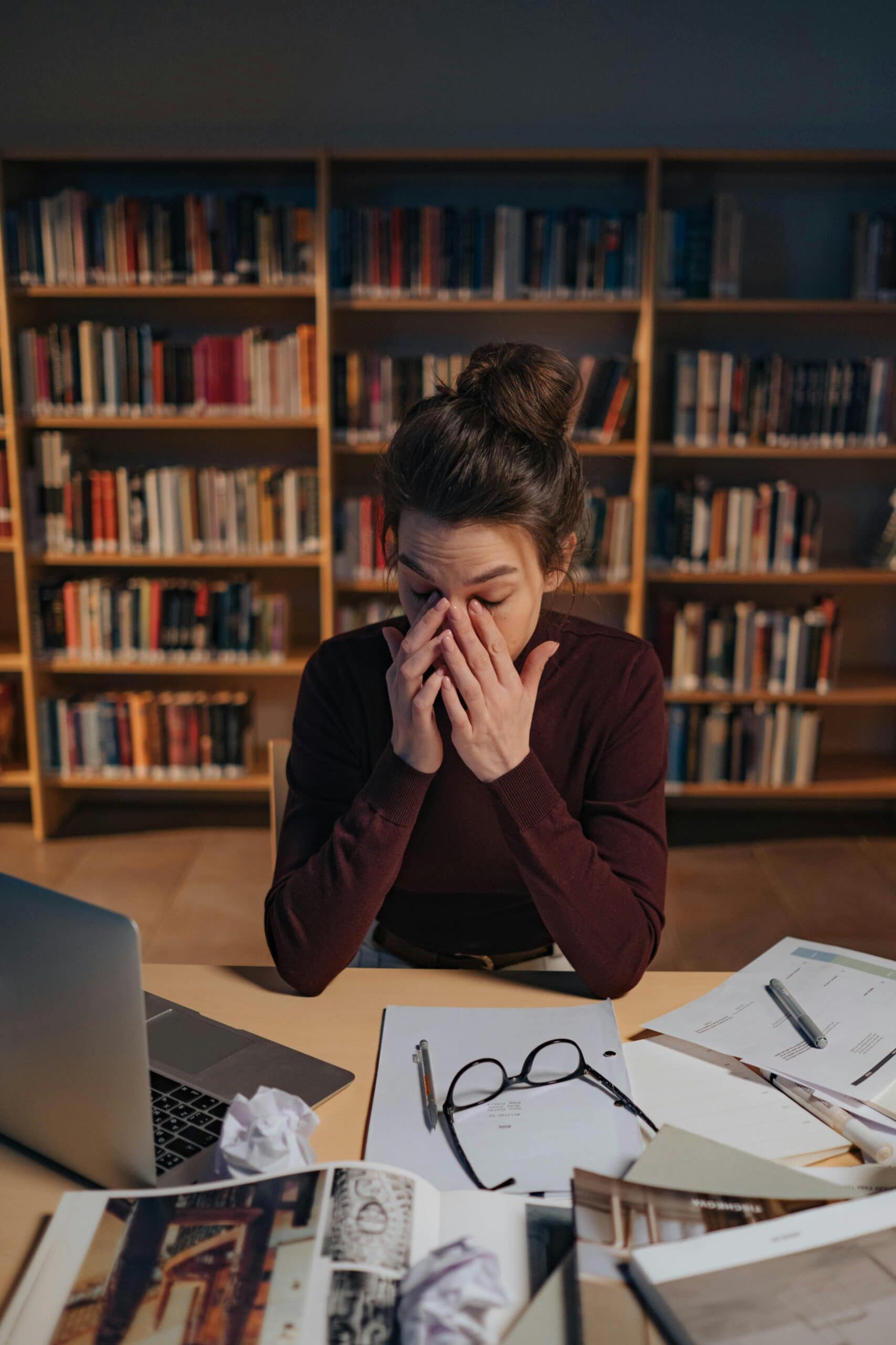 Student sitting at a library desk rubbing her eyes from stress
